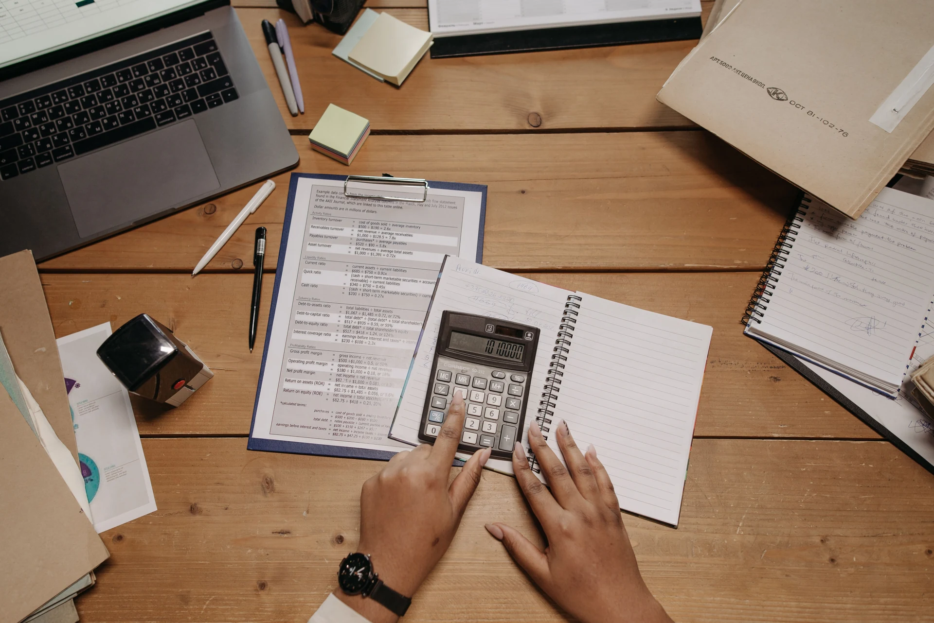 Plano cenital horizontal, manos en calculadora, documentos en carpeta, cuaderno y computador; sin planos mecánicos.